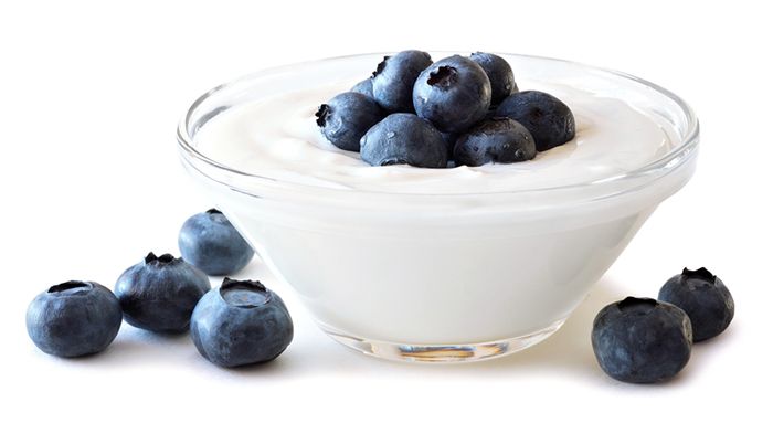 Clear small glass bowl filled yoghurt, blueberries on both sides of the bowl and some on the yoghurt. White isolated background